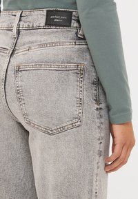 Vaqueros de denim de color gris claro con cintura alta, que presentan una etiqueta negra en la parte trasera y bolsillos standard. La textura está ligeramente desgastada.
