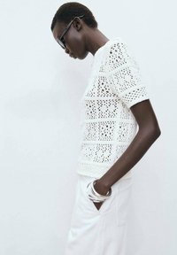White crocheted short-sleeve top with openwork pattern; model wears silver bangles and white pants; styled with black sunglasses.