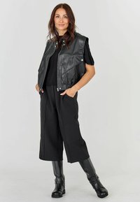 Hosszú barna hajú nő fekete bőrmellettes, fekete rövid szárú, széles lábú nadrágban, fekete rövid ujjú pólóban és fekete térdig érő csizmában áll, kezét a zsebében tartva.