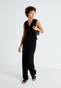 Gilet sans manches noir à deux boutons, associé à un pantalon large noir. Porté avec des sandales à talons noires et un petit sac à main noir.