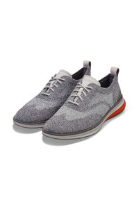 Zapatillas de punto gris con un patrón texturizado, que cuentan con un cierre de cordones blanco, suela gris y un toque de naranja en el talón.