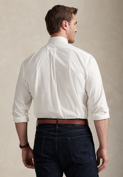 Man wearing white long-sleeve shirt with sleeves rolled up, dark jeans, and brown leather belt, viewed from the back against neutral background.