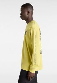 Long-sleeve yellow shirt featuring a logo on the chest and text along the sleeve. The fabric is smooth with a relaxed fit.