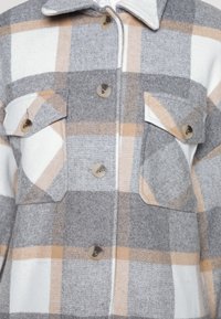 Camisa de cuadros abotonada hecha de suave lana gris y beige. Cuenta con dos bolsillos en el pecho, cuello redondeado y grandes botones claros.