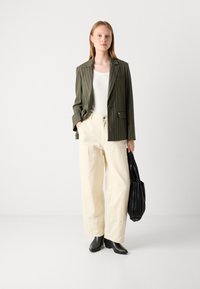 Oliivroheline pinstripe bleiser, valge pluus, kreemjad laiad püksid ja mustad pahkluu saapad. Hoides musta volditud kotti. Lihtne taust.