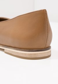 Gros plan sur le talon d'une chaussure plate en cuir marron avec des coutures visibles et une semelle à plusieurs couches aux tons beige et marron sur une surface blanche.