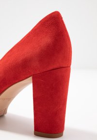 Escarpin en daim rouge à talon haut, bout pointu et talon épais. La texture est douce et mate, avec une semelle lisse de couleur marron.