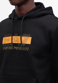 Άνδρας που φοράει μαύρο φούτερ με κουκούλα με το λογότυπο EA7 Emporio Armani σε κίτρινο και πορτοκαλί στο στήθος, με κορδόνια και τσέπη μπροστά.