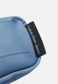 Blå texturerad tygpåse med rundade kanter och en svart etikett med "Calvin Klein Jeans" i vit text på en böjd yta.