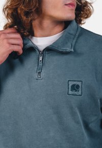 Pullover in pile, colore teal, design a zip a un quarto, texture liscia, con una toppa quadrata con logo di un elefante sul lato sinistro del petto.