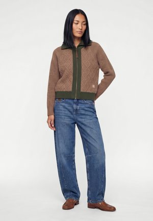 Kvinde med glat sort hår iført en brun mønstret sweatjakke med lynlås, løse blå jeans og brune sko, stående mod en ensfarvet baggrund.