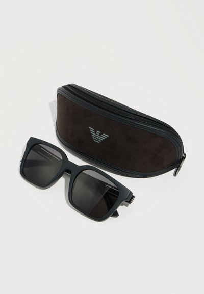 Μαύρα ορθογώνια γυαλιά ηλίου με σκούρους φακούς δίπλα σε μια μαύρη θήκη με φερμουάρ που φέρει ασημένιο λογότυπο αετού Armani σε λευκό φόντο.