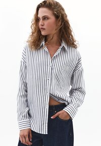 Donna con capelli ricci, che indossa una camicia bianca con bottoni e strisce verticali nere, jeans blu scuro, con una mano in tasca, che guarda a sinistra.