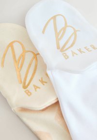 Divas divas zeķu pāri, viens pasteļbrūns ar zelta logo, otrs pilnīgi balts ar zelta logo, abi piedāvā mīkstu tekstūru un gludu apdari.