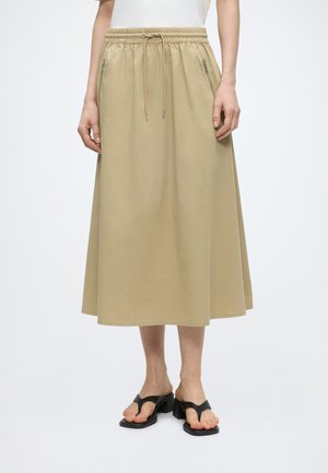 Jupe midi beige avec taille élastique et cordon, poches latérales zippées, portée avec des sandales à talons noires et un haut blanc.