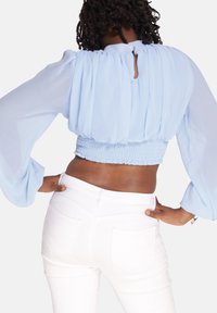 Jasnoniebieska krótka bluzka z przezroczystej tkaniny z bufkami, marszczonym stanem i zapięciem na kluczyk z tyłu. W zestawieniu z białymi dżinsami.