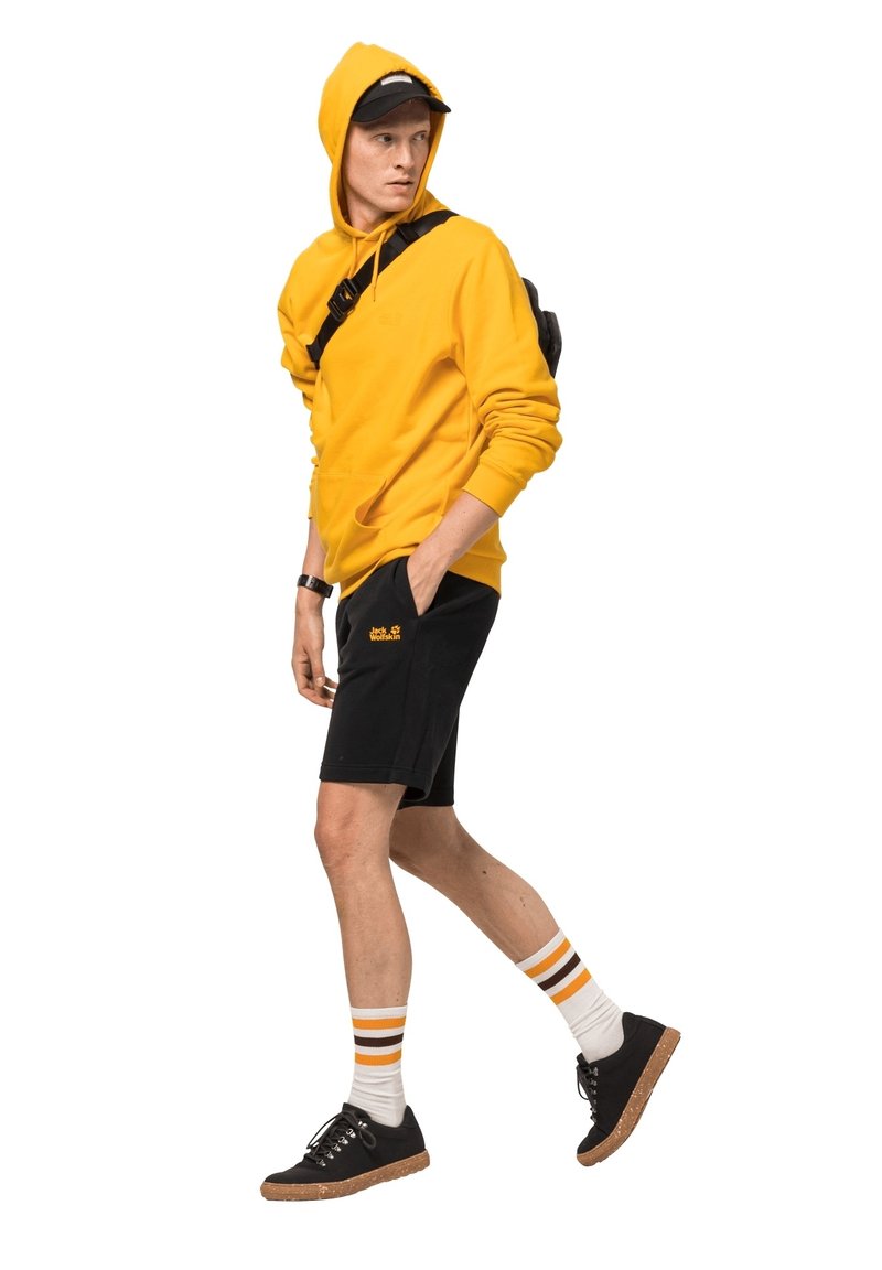 Gele hoodie, zwarte shorts met logo, wit en oranje gestreepte sokken, zwarte sneakers met kurkenzolen. Casual streetwear stijl.