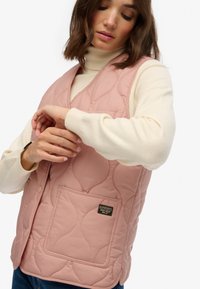 Gilet matelassé rose avec un col en V, doté d'une poche sur le devant et d'une texture lisse, superposé à un pull en col roulé crème.