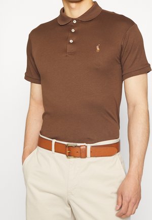 Polo marrone con collo classico e patta con tre bottoni; presenta un logo ricamato sul petto. Abbinato a pantaloni beige chiaro e cintura in pelle.