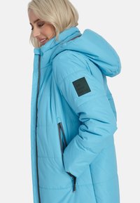 Gaiši zils pūkains jaka ar kapuci, ar ieņemamu kabatu un auduma uzlīmi uz piedurknes ar tekstu "URBAN WEAR". Gluda tekstūra.