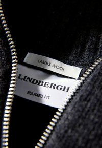 Svart lammull-klädesetikett med vit text som säger "LINDENBERGH," "AVSLAPPNAD PASSFORM," och "LAMMSULL," broderad nära en silverdragkedja.