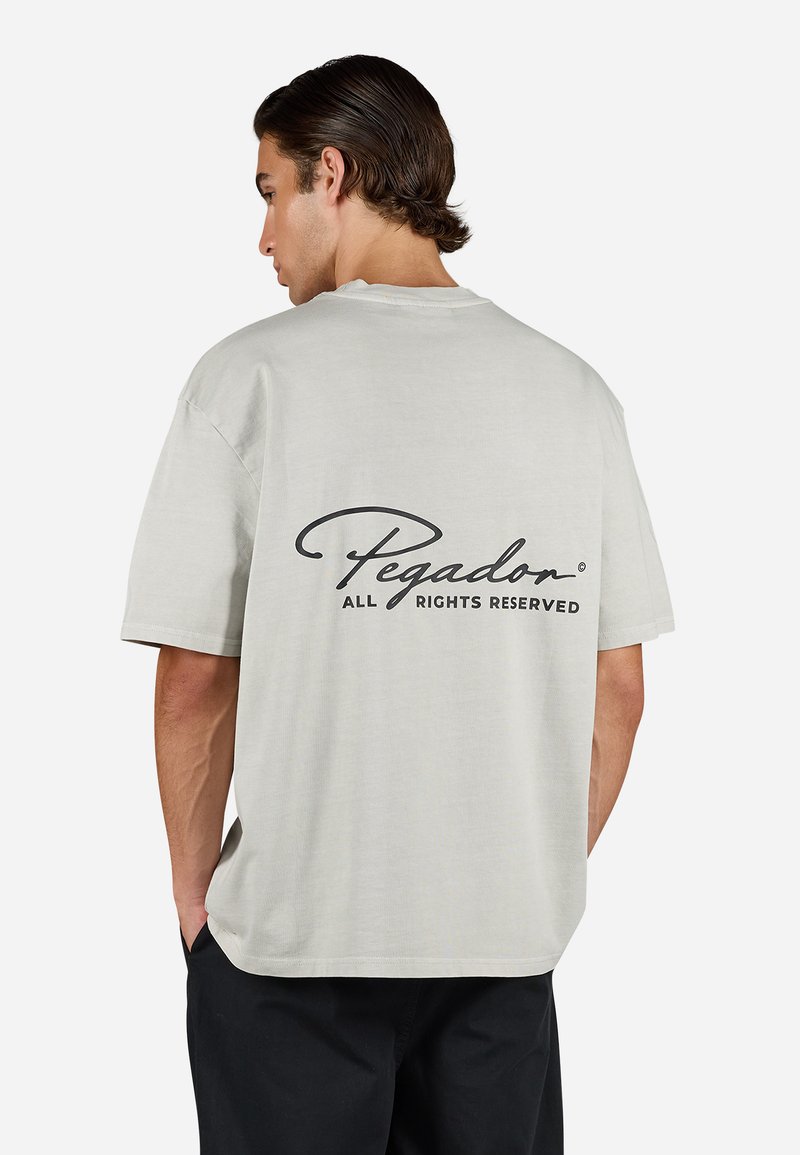Lys grå oversized t-shirt med korte ærmer. Har et sort trykt logo med teksten "Pegador" på ryggen. Blødt bomuldsmateriale.