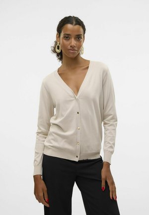 Cardigan beige avec une texture douce, décolleté en V et détails de boutons dorés. Manches longues et coupe ajustée ; porté avec un pantalon noir.