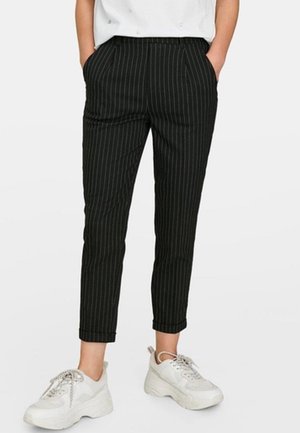 Persoon draagt zwarte cropped broek met krijtstrepen en opgerolde zomen, gecombineerd met witte chunky sneakers, handen in de zakken, staand tegen een effen achtergrond.