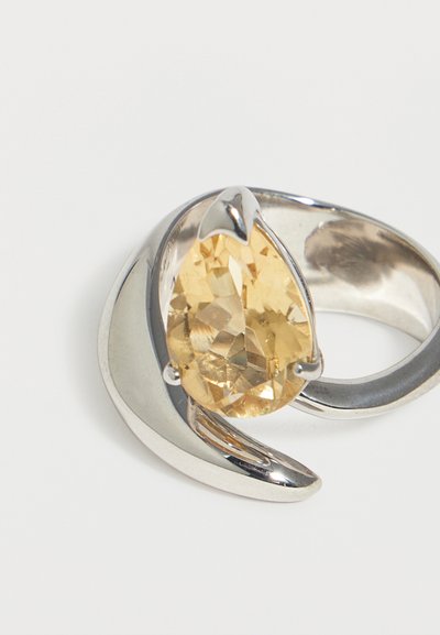 Ασημένιο δαχτυλίδι με μεγάλο αχλαδοειδές κίτρινο πολύτιμο λίθο, τοποθετημένο σε μοντέρνο, καμπυλωτό μεταλλικό σχέδιο σε λευκό φόντο.