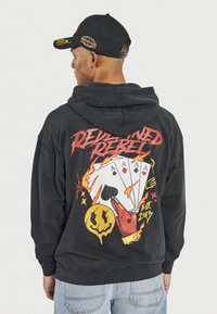 Schwarzer Hoodie mit einem großen Grafikdesign von Spielkarten, Flammen und einem Smiley-Gesicht. Akzentuiert mit roten und gelben Farben. Aus Baumwollmischung.