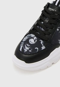 Čierne tenisky s textúrovanou koženou pätou, kontrastnou bielou podrážkou a dekoratívnym čierno-bielym kvetinovým motívom na látke.