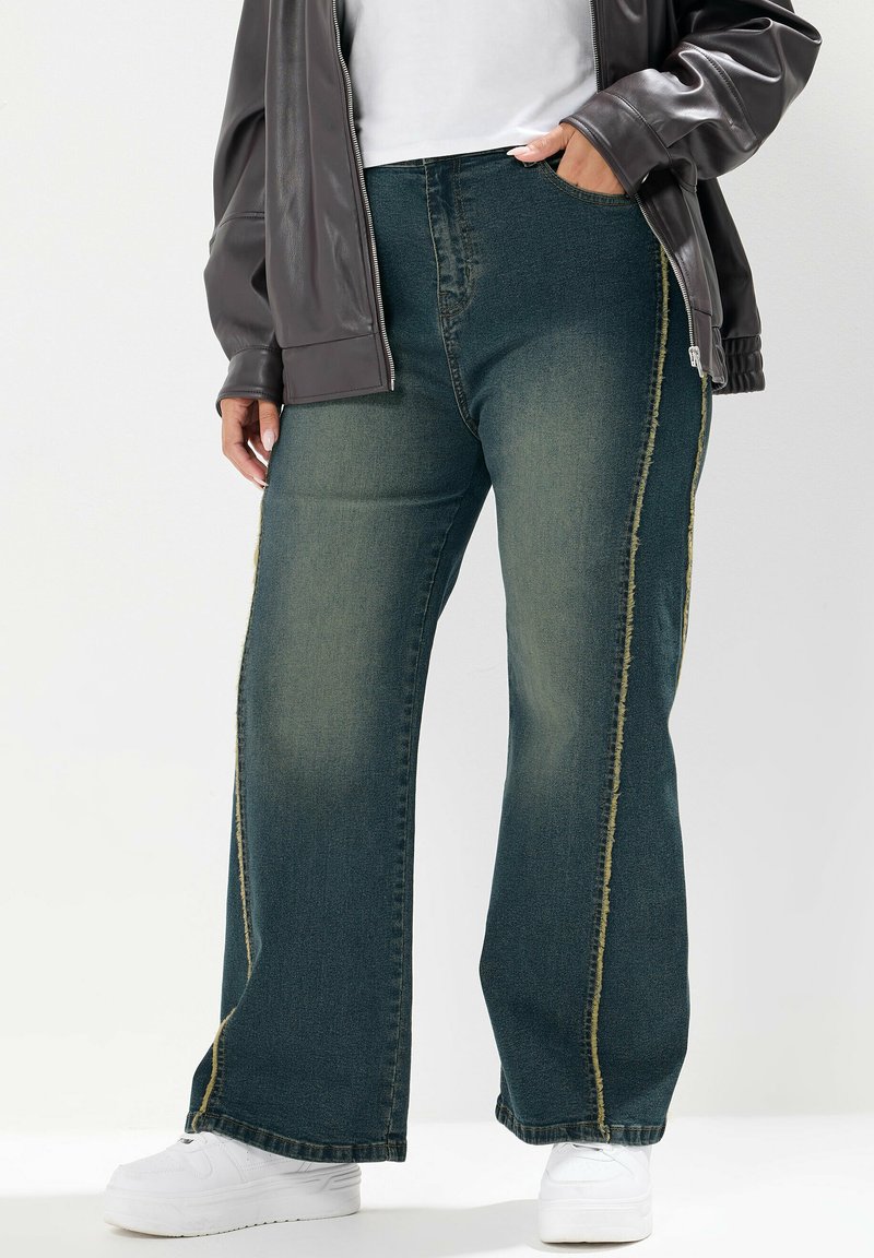 Väldigt vida, mörkgröna denimjeans med ljusa slitningar och gula sömmar längs sidsömmarna. I kombination med en grå jacka och vita skor.