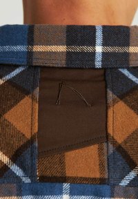 Geruiten flanellen shirtkraag met een blauw, bruin en wit ruitpatroon; de stof lijkt zacht met een patch en stiksel detail aan de achterkant.