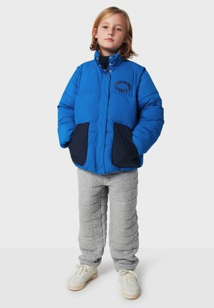 Blå pufferjakke med høj krave, sorte lommer og logo detaljer. Sammen med grå quiltede bukser og hvide sneakers.