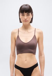 Γυναίκα με κοντά σκούρα μαλλιά, φορώντας καφέ cropped τοπ και μαύρο ριμπ ύφασμα εσώρουχο με ψηλό κόψιμο, στέκεται μπροστά σε ένα απλό φωτεινό φόντο.