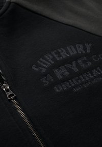 Zwarte hoodie met rits, featuring een getextureerd geborduurd logo, gladde stof en een zilveren rits, met contrasterende donkergroene bovenmouwen.