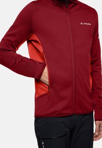 Rote und orange Zip-Jacke mit langen Ärmeln, strukturiertem Stoff und seitlichen Taschen mit Reißverschluss. Markenlogo auf der Brust.