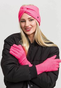 Bandeau en tricot rose avec un design tordu, associé à des gants côtes roses assortis. Le modèle porte une doudoune noire.
