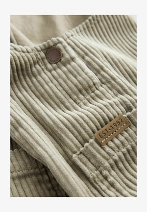 Tejkrém színű kordbársony anyag függőleges bordázással, amelyen egy barna kör alakú gomb és egy "EST-1982" feliratozott téglalap alakú címke található.