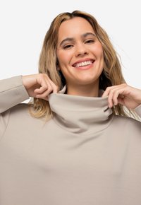 Suéter de cuello alto beige con una textura suave y elástica, de corte holgado y con cuello doblado. Modelo tirando suavemente del escote.