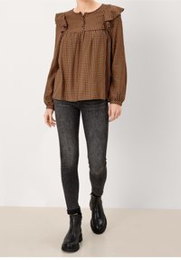 Blusa a scacchi marrone e nera con dettagli arricciati, scollatura rotonda, dettagli con bottoni e maniche lunghe. Abbinata a jeans skinny grigi scuri e stivali neri.
