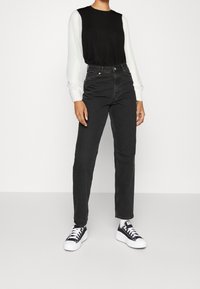 Persona che indossa jeans neri a vita alta, top nero con maniche lunghe bianche testurizzate, sneakers in bianco e nero, in piedi contro uno sfondo chiaro e semplice.
