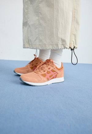 Personne portant un pantalon large beige, des chaussettes blanches et des baskets couleur pêche, debout sur un tapis bleu clair.
