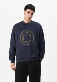 Sudadera azul marino con un diseño redondo de alambre de espino que presenta una cara sonriente y las letras "HGO" en bordado dorado. Puños y dobladillo acanalados.