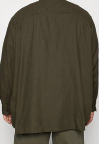 Camicia verde oliva a maniche lunghe realizzata in un tessuto strutturato. Presenta un taglio dritto, polsini con bottone e un dettaglio sullo yoke posteriore.