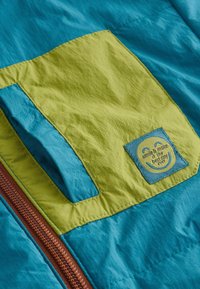 Sección de chaqueta colorida con paneles en color azul verdoso y lima. Incluye un zipper marrón y una etiqueta cosida con un motivo de cara sonriente.