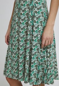 Robe florale en tissu léger, avec un motif floral vert et noir agrémenté de touches de jaune. Forme en A et longueur au genou.