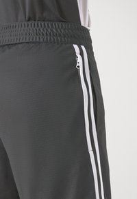 Gros plan sur un short de sport gris foncé avec une ceinture élastique et des rayures blanches verticales à côté d'une poche latérale zippée.