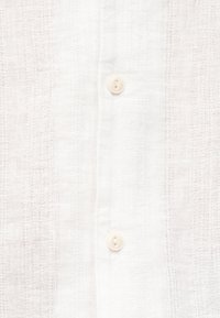 Κοντινή λήψη μπροστινής όψης λευκού λινό πουκάμισου με κάθετη υφή ύφανσης και δύο μικρά στρογγυλά λευκά κουμπιά.