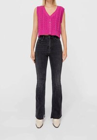 Femme portant un gilet en maille sans manches rose vif, un jean en denim noir taille haute avec des fentes à l'avant, et des chaussures pointues couleur crème.